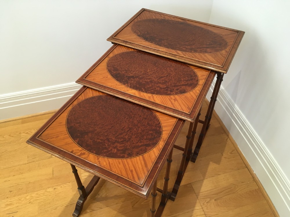 A set of three nested wooden side tables featuring rectangular tops with oval amboyna wood centres, inlaid satinwood borders, and slender turned legs.
