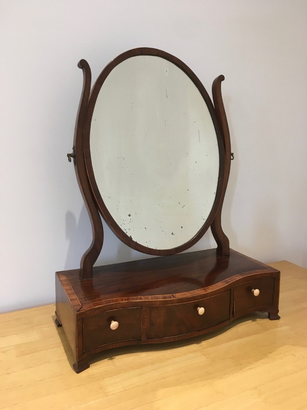An antique Georgian mahogany toilet mirror with an oval pivoting mirror above a serpentine base fitted with three small drawers and light-coloured knobs.