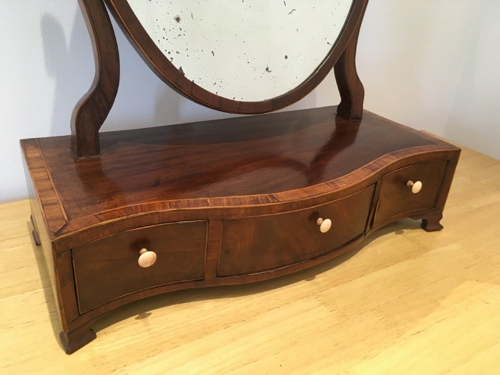 An antique Georgian mahogany toilet mirror with an oval pivoting mirror above a serpentine base fitted with three small drawers and light-coloured knobs. View II