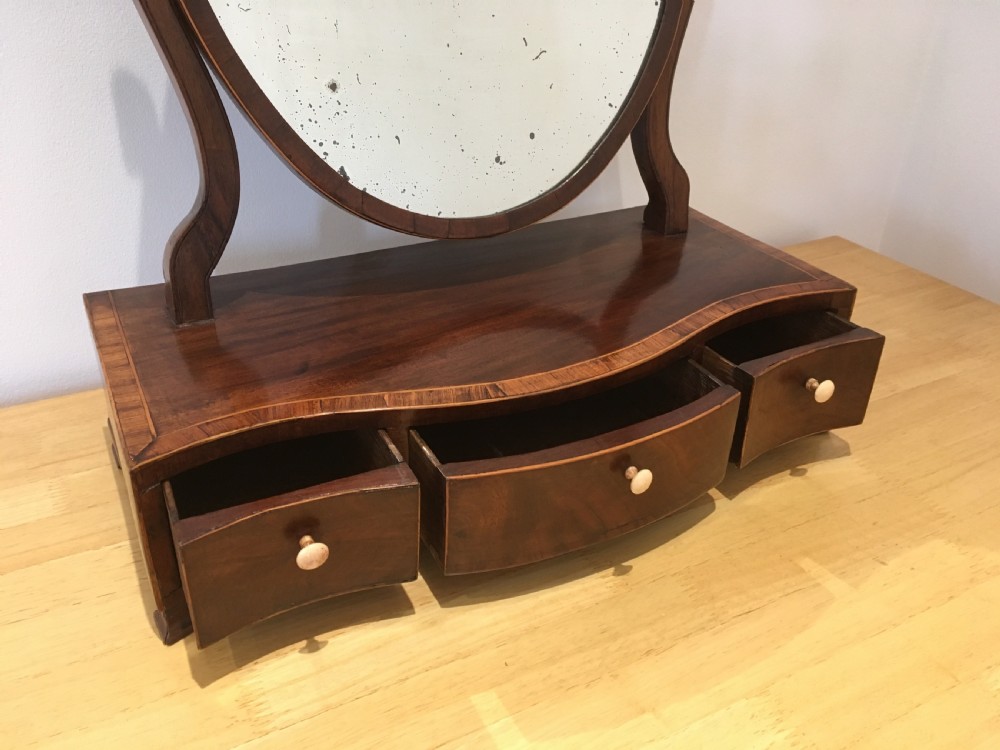 An antique Georgian mahogany toilet mirror with an oval pivoting mirror above a serpentine base fitted with three small drawers and light-coloured knobs. View III