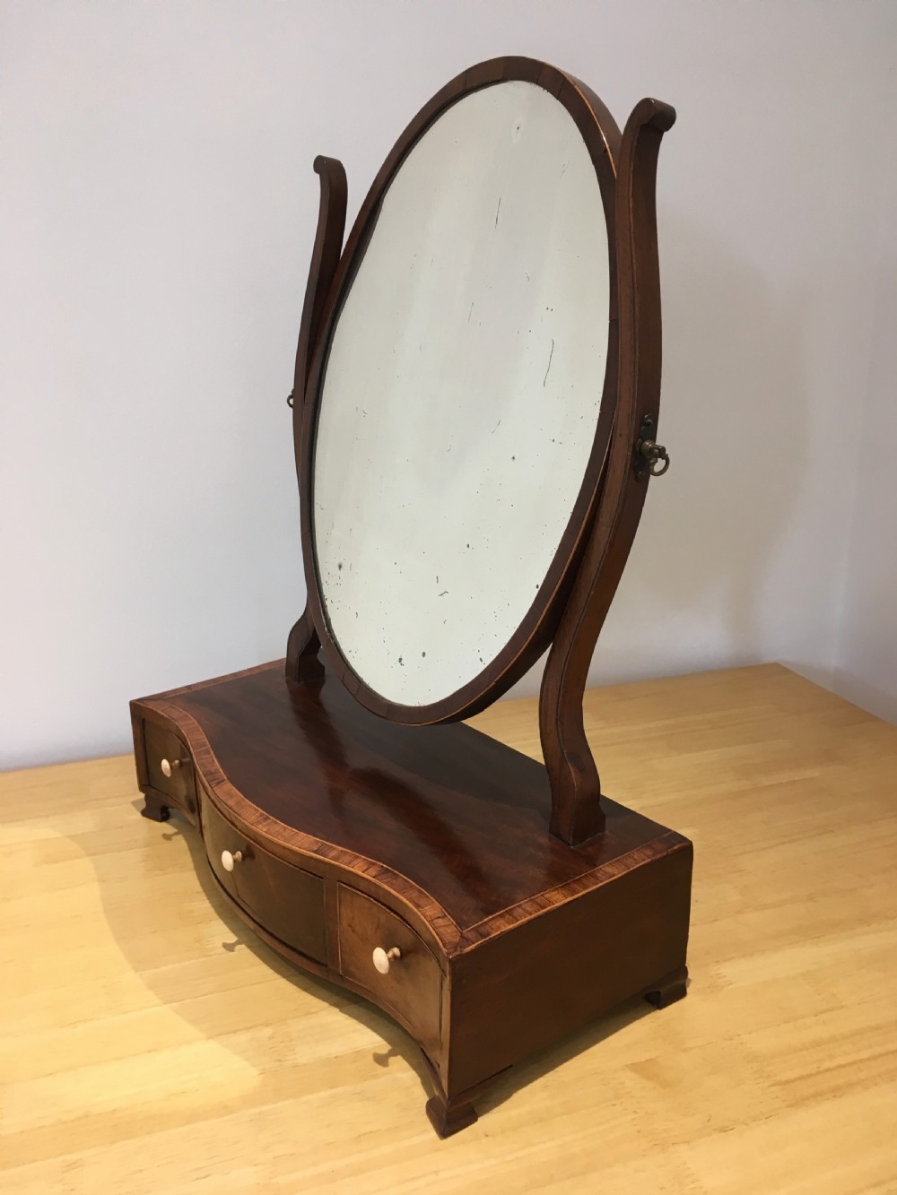 An antique Georgian mahogany toilet mirror with an oval pivoting mirror above a serpentine base fitted with three small drawers and light-coloured knobs. View IV
