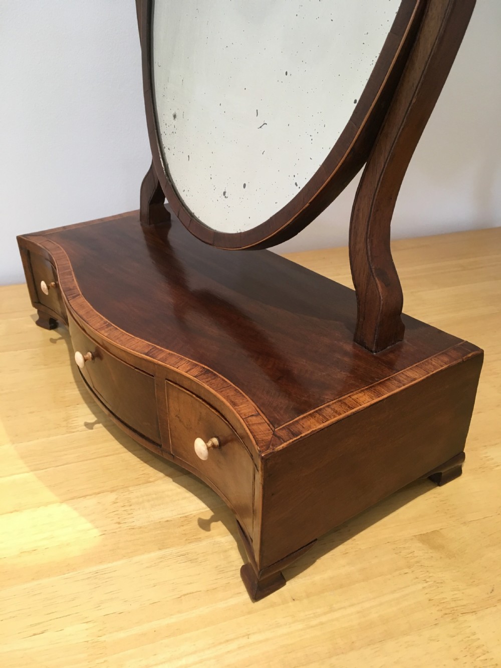 An antique Georgian mahogany toilet mirror with an oval pivoting mirror above a serpentine base fitted with three small drawers and light-coloured knobs. View V