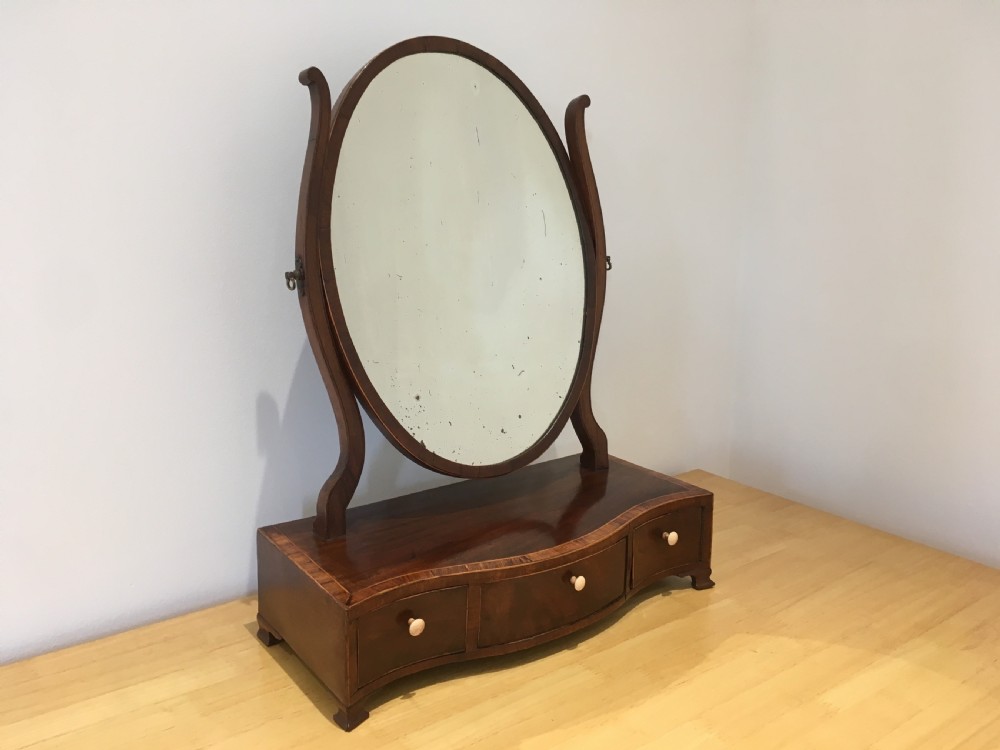 An antique Georgian mahogany toilet mirror with an oval pivoting mirror above a serpentine base fitted with three small drawers and light-coloured knobs. View VII
