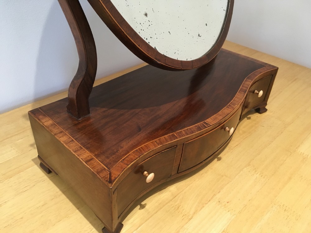 An antique Georgian mahogany toilet mirror with an oval pivoting mirror above a serpentine base fitted with three small drawers and light-coloured knobs. View IX