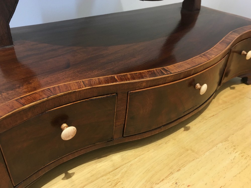 An antique Georgian mahogany toilet mirror with an oval pivoting mirror above a serpentine base fitted with three small drawers and light-coloured knobs. View X