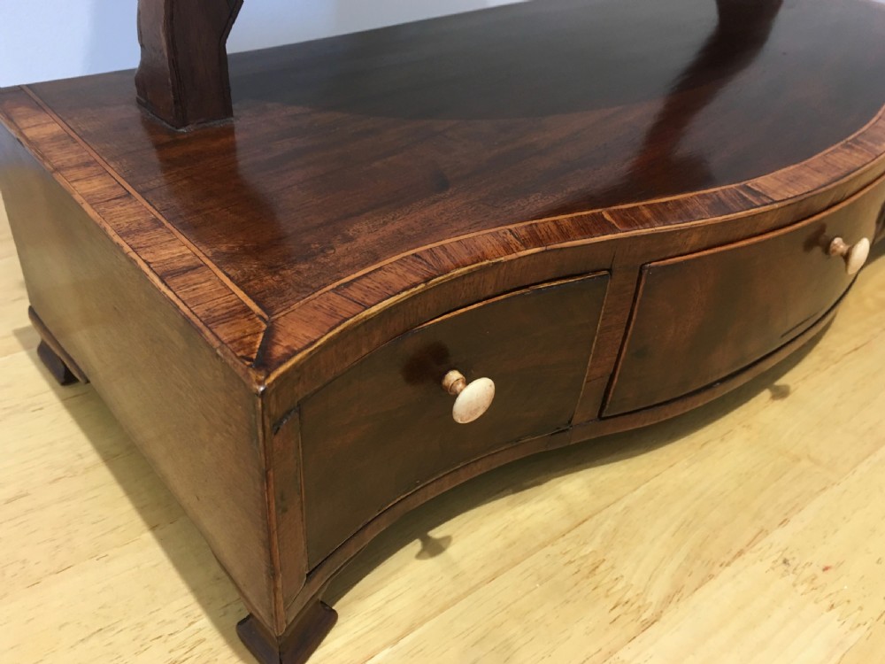 An antique Georgian mahogany toilet mirror with an oval pivoting mirror above a serpentine base fitted with three small drawers and light-coloured knobs. View XI