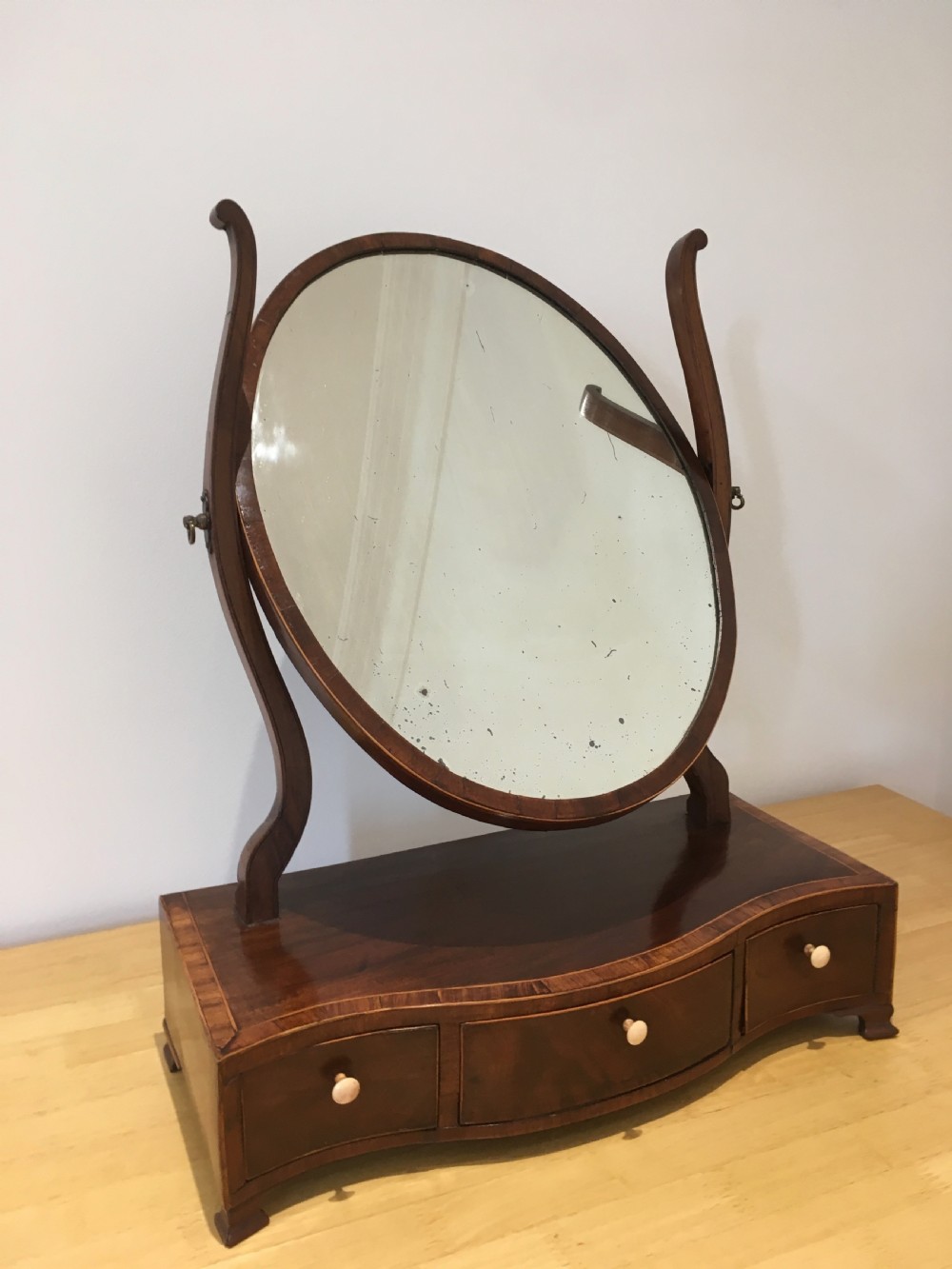 An antique Georgian mahogany toilet mirror with an oval pivoting mirror above a serpentine base fitted with three small drawers and light-coloured knobs. View XII