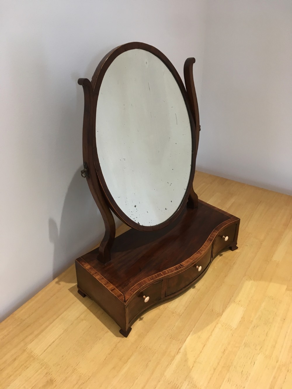 An antique Georgian mahogany toilet mirror with an oval pivoting mirror above a serpentine base fitted with three small drawers and light-coloured knobs. View XIII