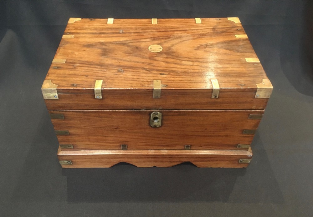 A rectangular warm brown oak desk box featuring brass bound corners, metal straps, a central brass oval cartouche, and a front keyhole escutcheon.