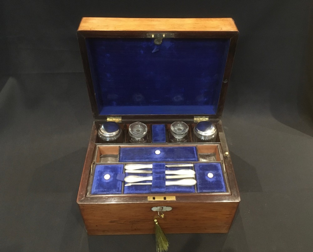 An open rosewood vanity box lined with vibrant blue velvet, containing cut glass jars, silver-plated lids, and manicure tools.