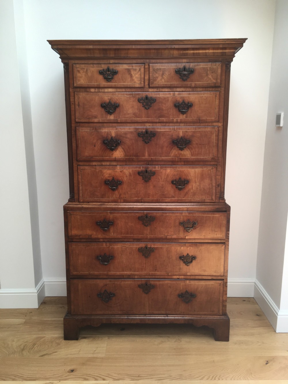 A George II walnut tallboy chest on chest featuring figured wood veneers, dark brass backplate handles, a moulded cornice, and bracket feet.