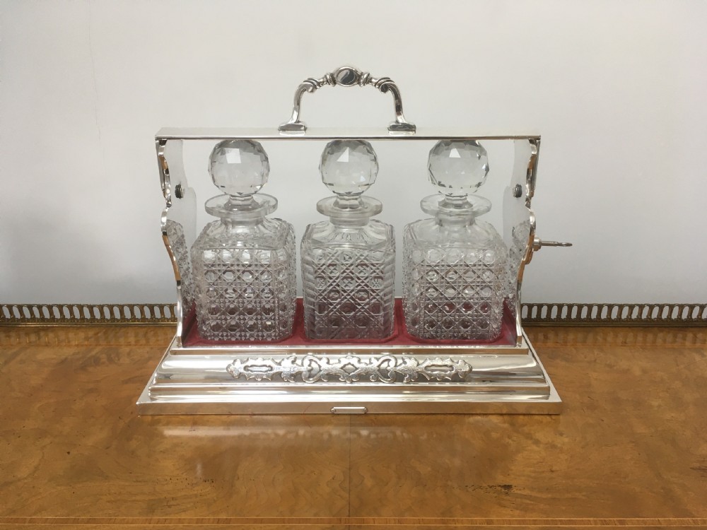 A silver-plated three-decanter tantalus featuring three cut-glass square decanters with facet-cut stoppers secured in a locking frame with a central handle.