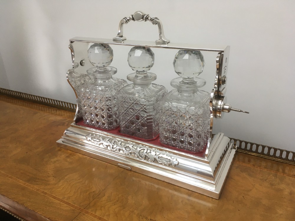 A silver-plated three-decanter tantalus featuring three cut-glass square decanters with facet-cut stoppers secured in a locking frame with a central handle. View II