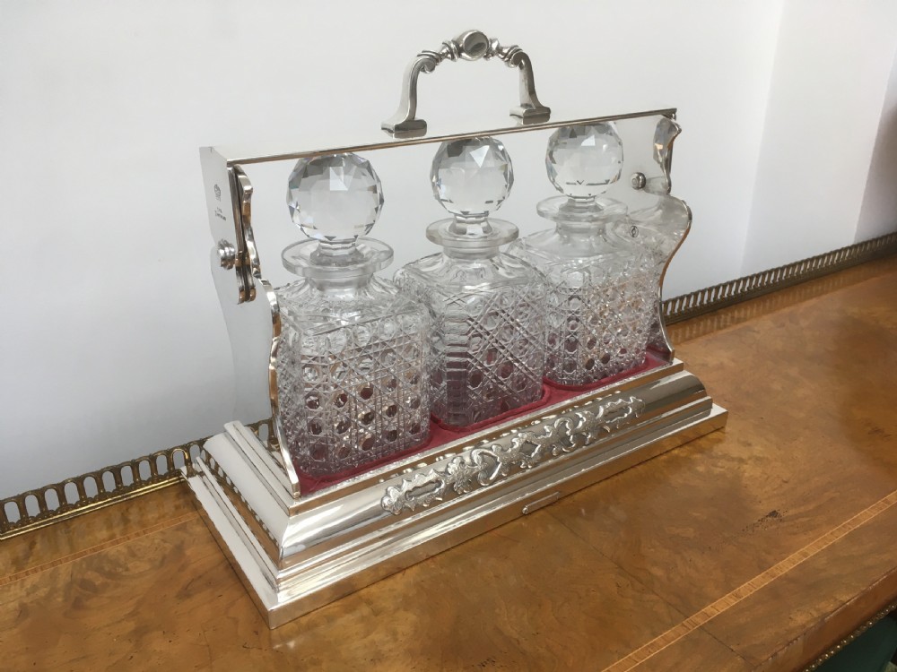 A silver-plated three-decanter tantalus featuring three cut-glass square decanters with facet-cut stoppers secured in a locking frame with a central handle. View IV