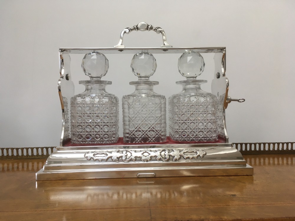 A silver-plated three-decanter tantalus featuring three cut-glass square decanters with facet-cut stoppers secured in a locking frame with a central handle. View VI