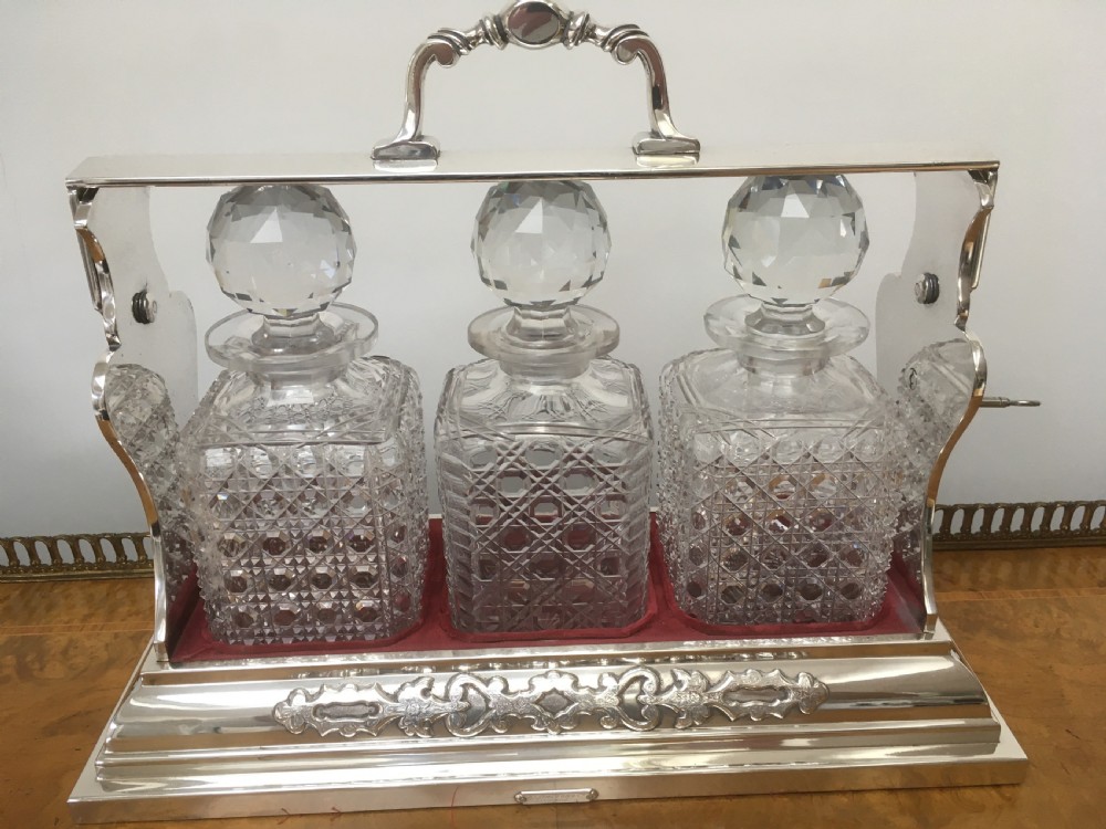 A silver-plated three-decanter tantalus featuring three cut-glass square decanters with facet-cut stoppers secured in a locking frame with a central handle. View IX