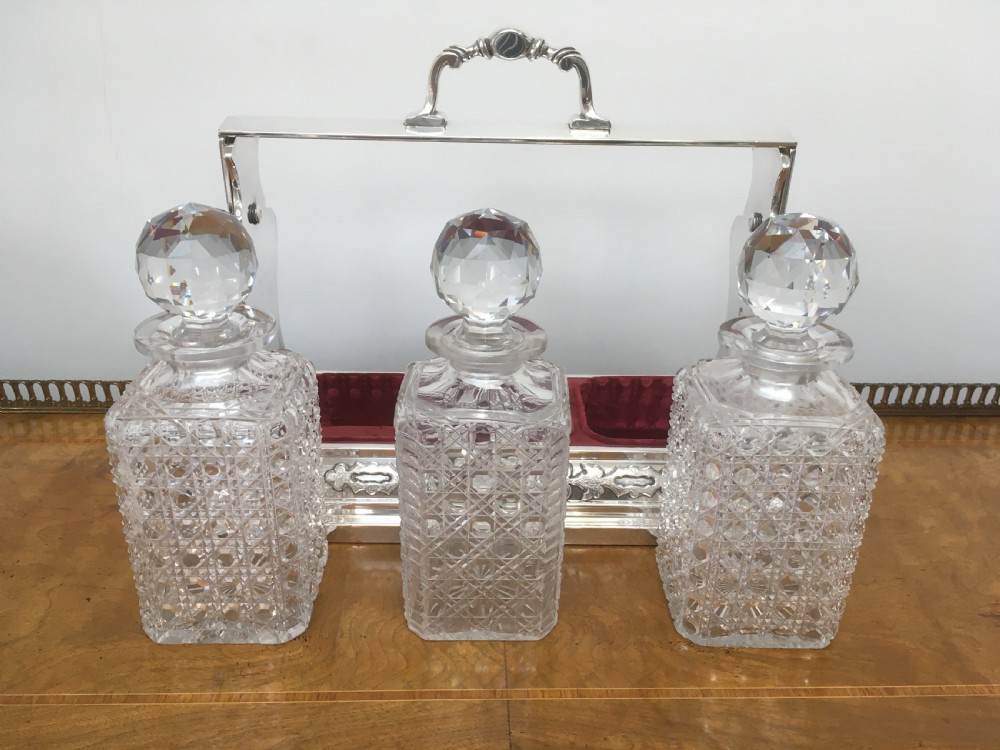 A silver-plated three-decanter tantalus featuring three cut-glass square decanters with facet-cut stoppers secured in a locking frame with a central handle. View X