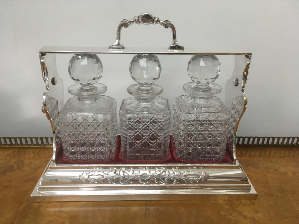 A silver-plated three-decanter tantalus featuring three cut-glass square decanters with facet-cut stoppers secured in a locking frame with a central handle. View XIV