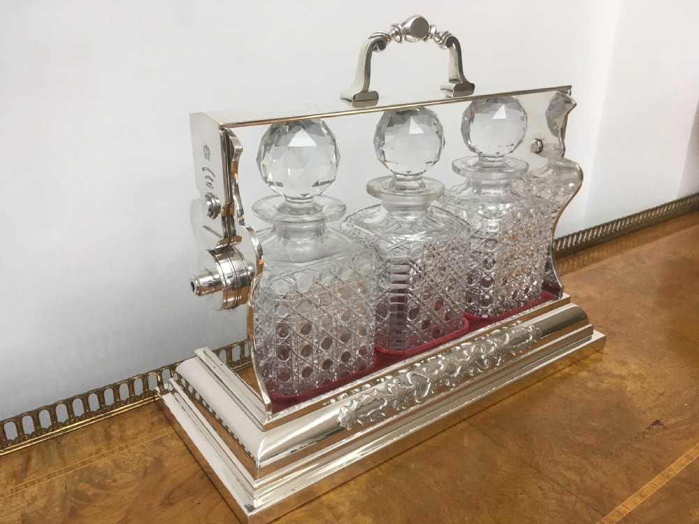 A silver-plated three-decanter tantalus featuring three cut-glass square decanters with facet-cut stoppers secured in a locking frame with a central handle. View XV