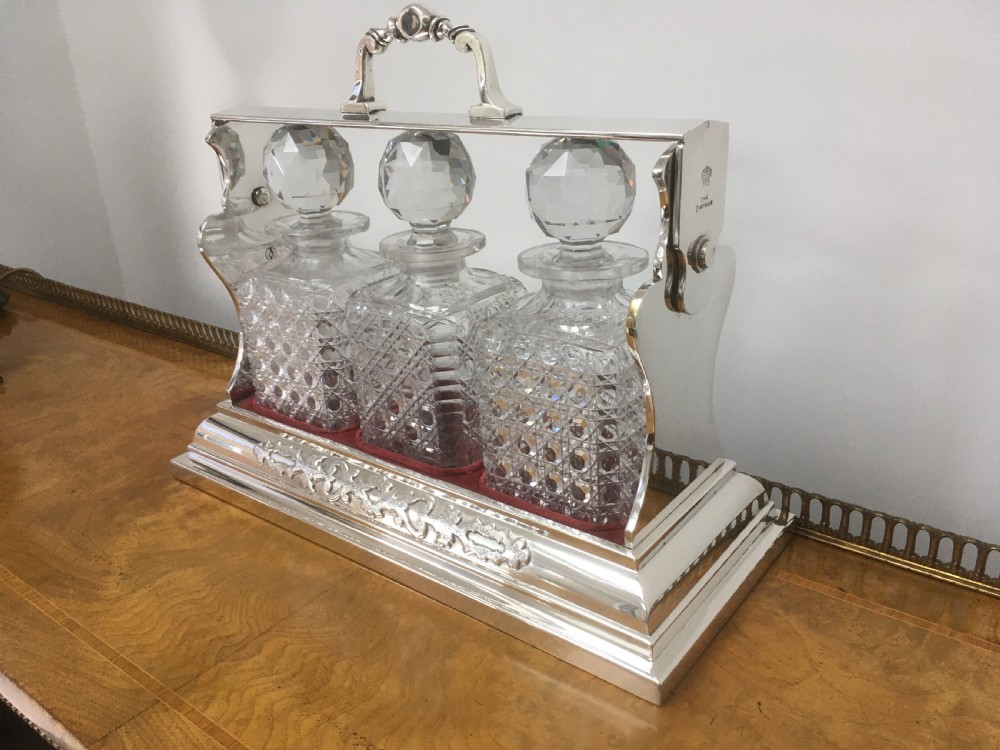 A silver-plated three-decanter tantalus featuring three cut-glass square decanters with facet-cut stoppers secured in a locking frame with a central handle. View XVI