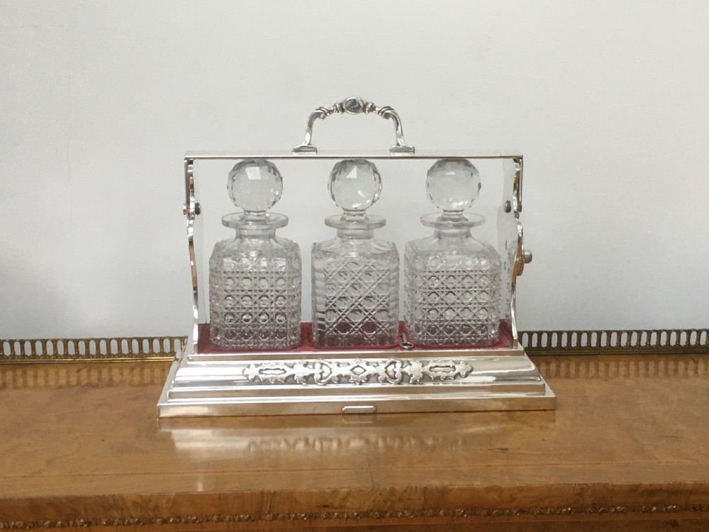 A silver-plated three-decanter tantalus featuring three cut-glass square decanters with facet-cut stoppers secured in a locking frame with a central handle. View XVII