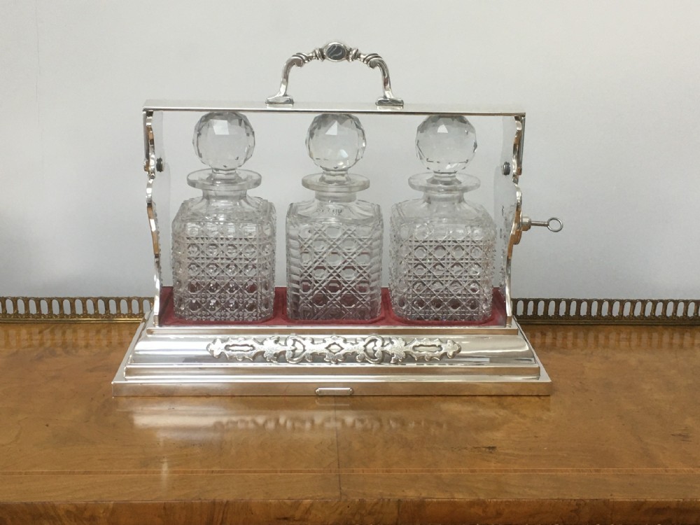 A silver-plated three-decanter tantalus featuring three cut-glass square decanters with facet-cut stoppers secured in a locking frame with a central handle. View XVIII
