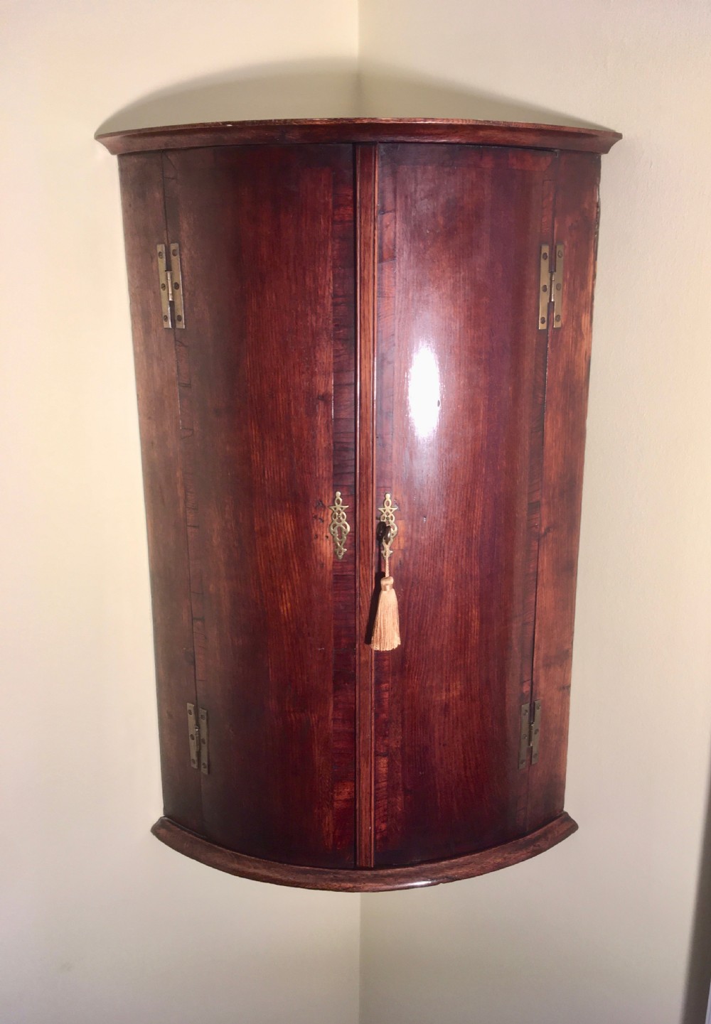 A wall-mounted Georgian corner cabinet featuring a curved mahogany bow front, brass H-hinges, and a central keyhole with a decorative tassel.