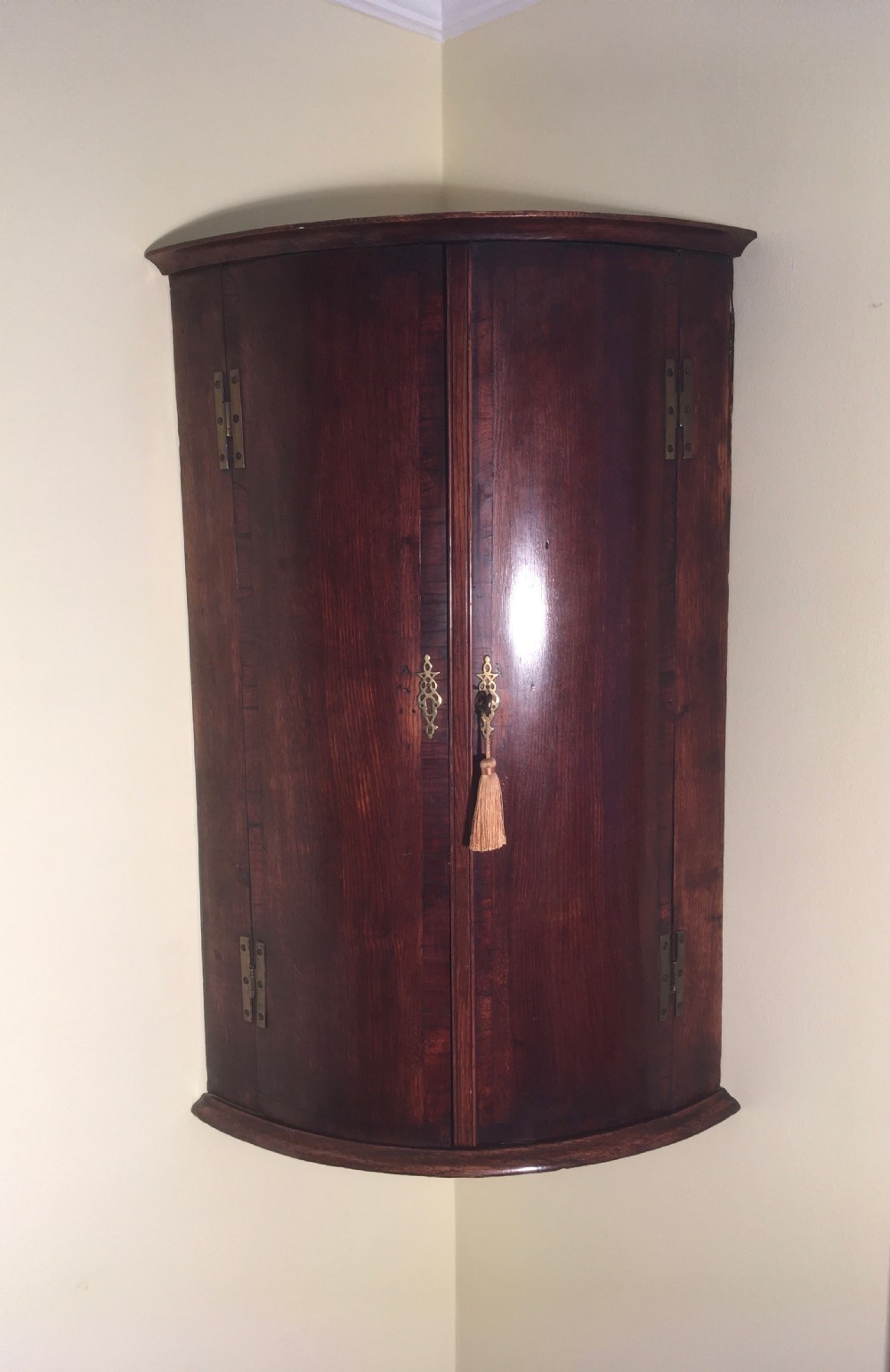 A wall-mounted Georgian corner cabinet featuring a curved mahogany bow front, brass H-hinges, and a central keyhole with a decorative tassel. View II