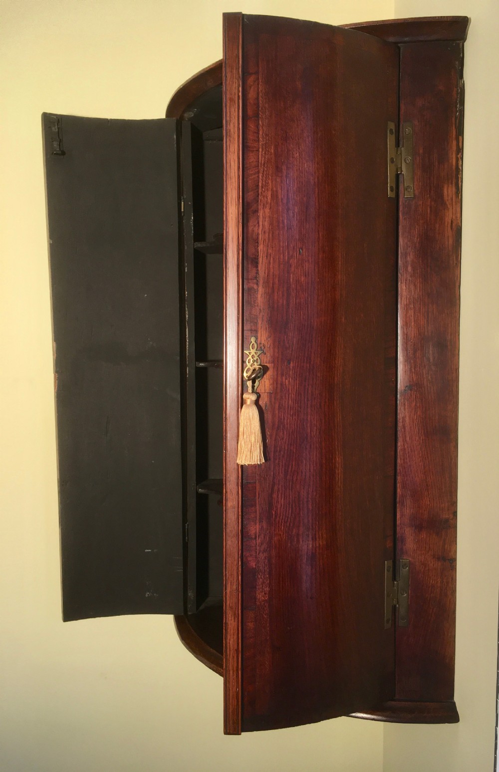 A wall-mounted Georgian corner cabinet featuring a curved mahogany bow front, brass H-hinges, and a central keyhole with a decorative tassel. View III