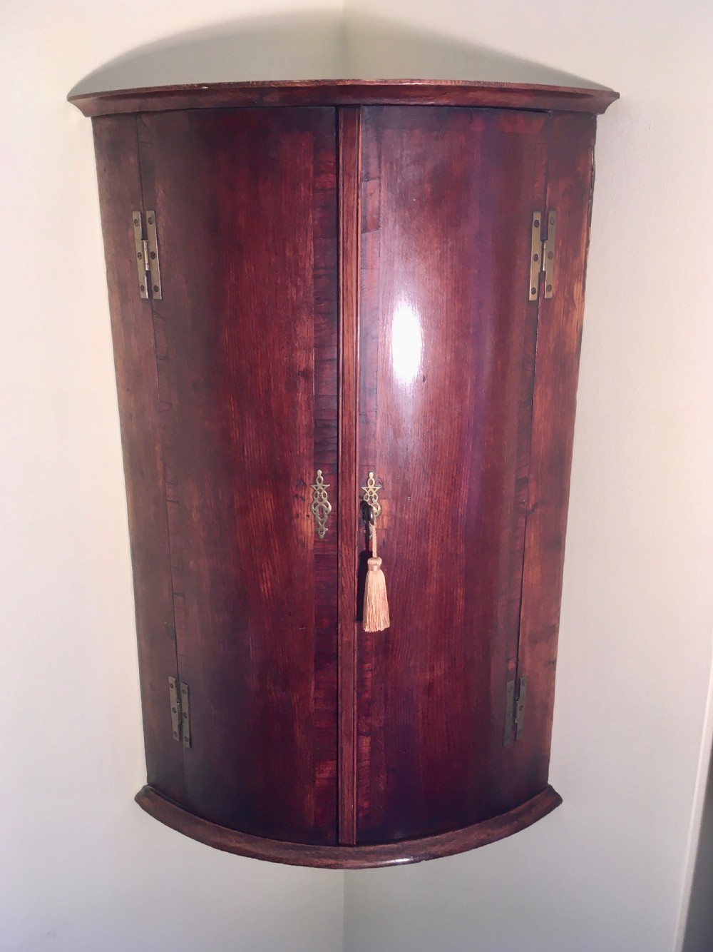 A wall-mounted Georgian corner cabinet featuring a curved mahogany bow front, brass H-hinges, and a central keyhole with a decorative tassel. View IX