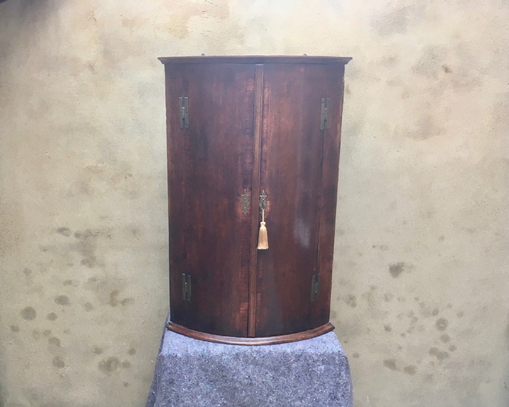 A wall-mounted Georgian corner cabinet featuring a curved mahogany bow front, brass H-hinges, and a central keyhole with a decorative tassel. View XIII
