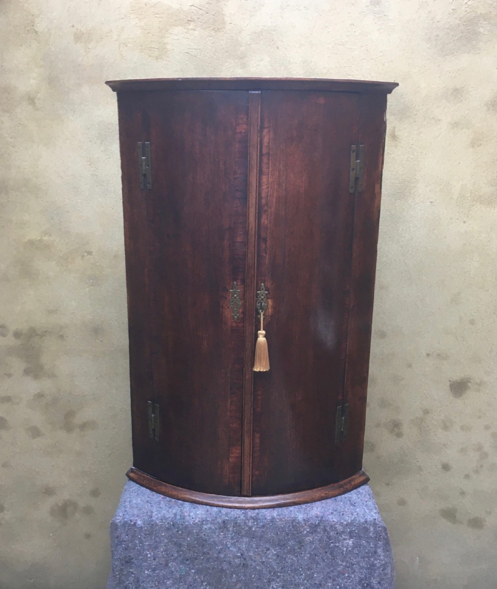 A wall-mounted Georgian corner cabinet featuring a curved mahogany bow front, brass H-hinges, and a central keyhole with a decorative tassel. View XIV