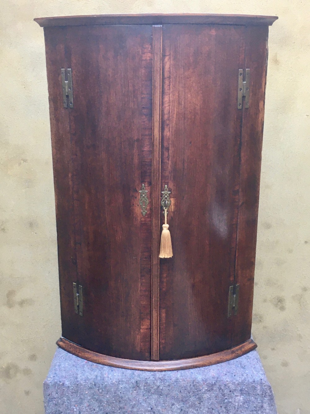A wall-mounted Georgian corner cabinet featuring a curved mahogany bow front, brass H-hinges, and a central keyhole with a decorative tassel. View XV
