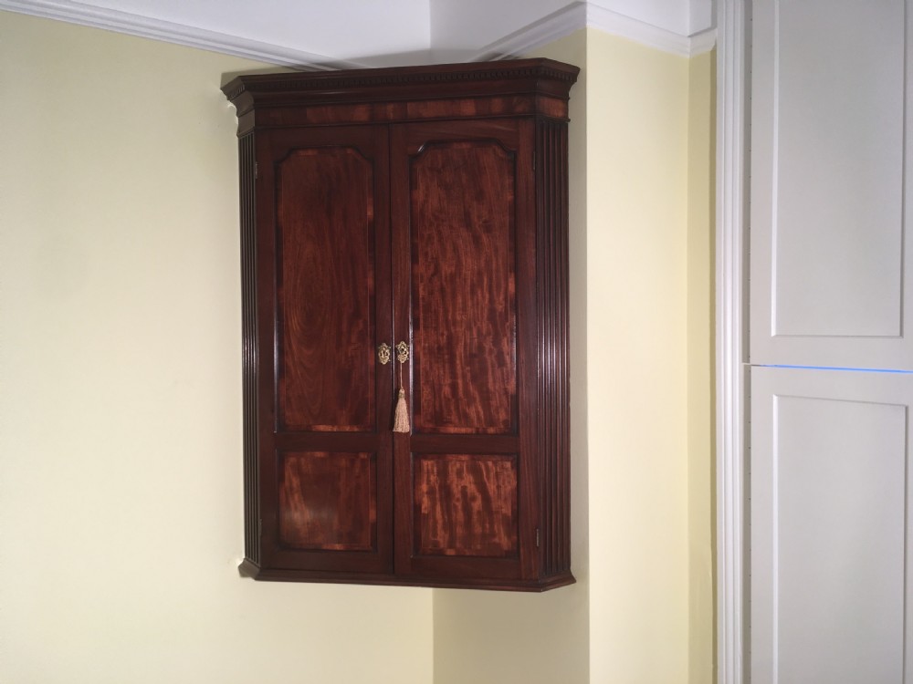A wall-mounted Georgian mahogany corner cabinet featuring twin panelled doors, fluted side pilasters, and brass keyhole escutcheons. View X