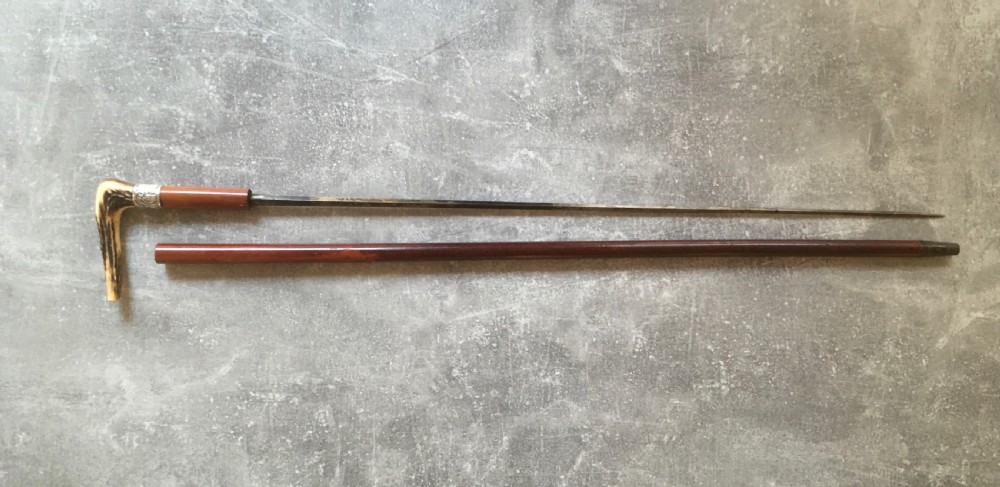 An antique Malacca wood walking stick featuring a carved stag antler handle and silver collar, drawn to reveal a long steel concealed blade. View II