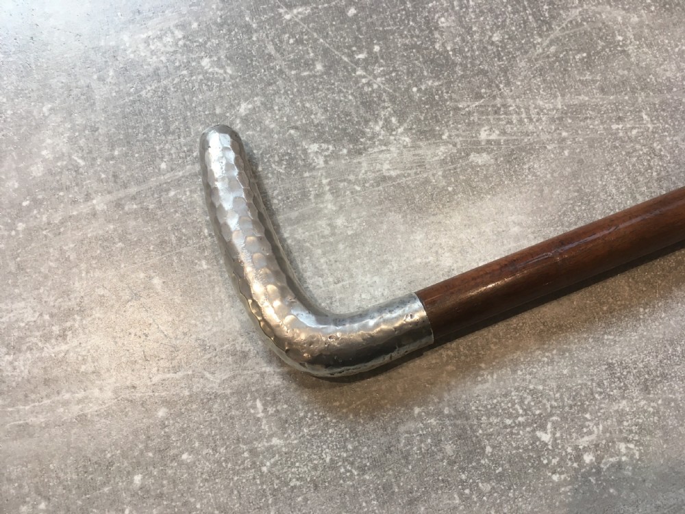 An antique walking stick featuring a hand-hammered silver L-shaped handle fitted to a tapered dark brown wooden shaft.