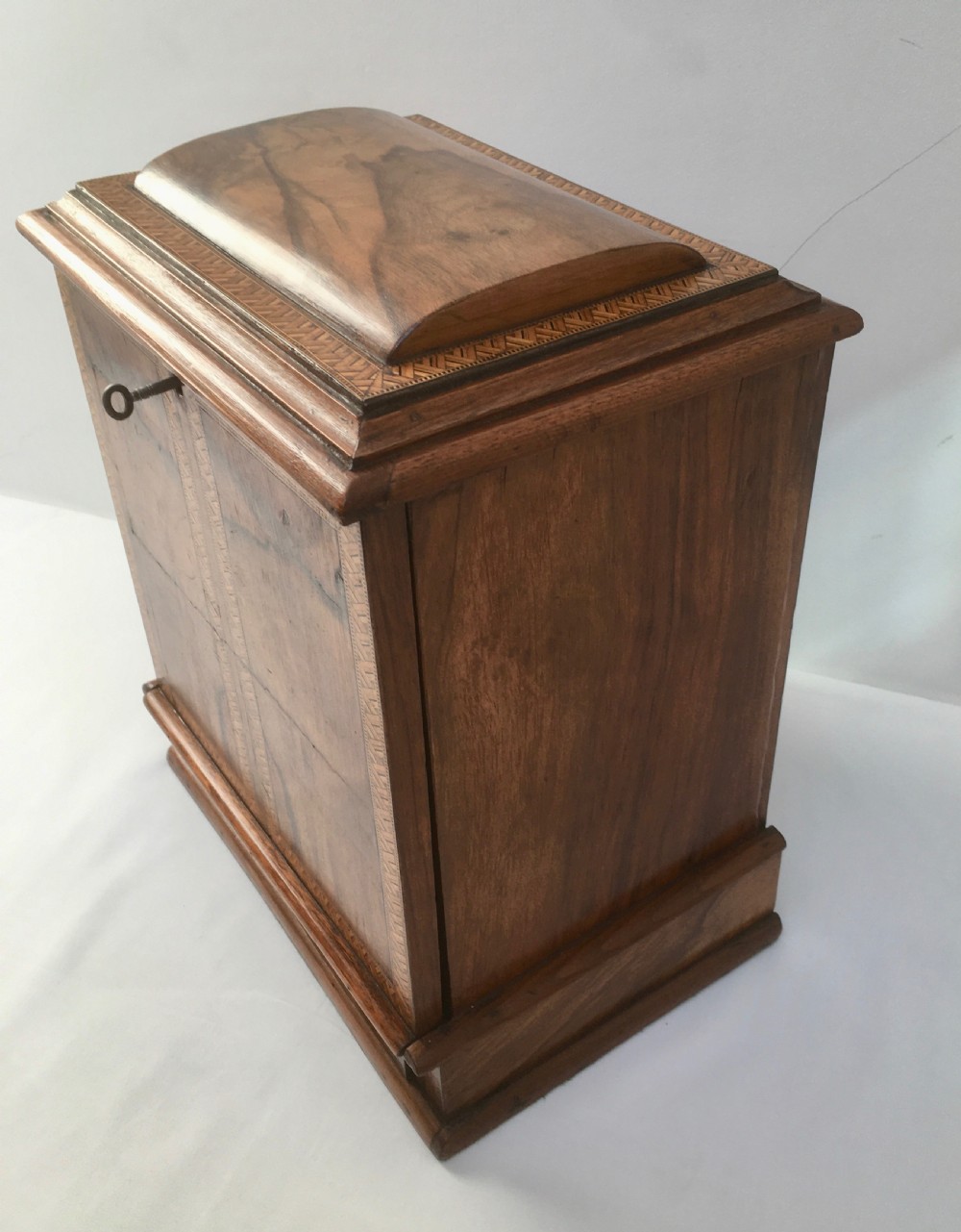 An inlaid walnut correspondence chest featuring geometric band inlay around figured wood panels, a domed lid, two front doors, and a central keyhole with a key inserted. View X