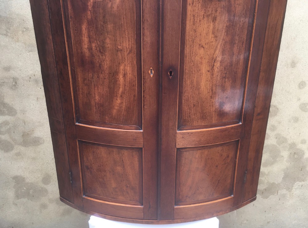 A Georgian mahogany bow-fronted corner cabinet featuring twin panelled doors, decorative inlay along the top cornice, and brass keyhole escutcheons. View X