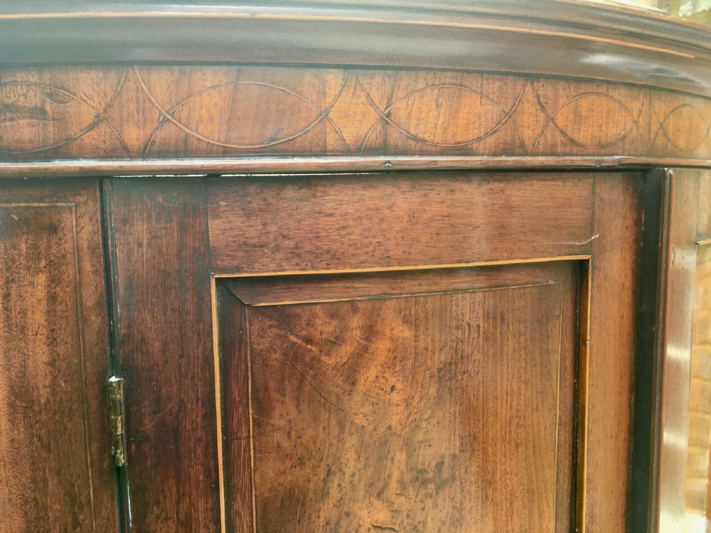 A Georgian mahogany bow-fronted corner cabinet featuring twin panelled doors, decorative inlay along the top cornice, and brass keyhole escutcheons. View XII