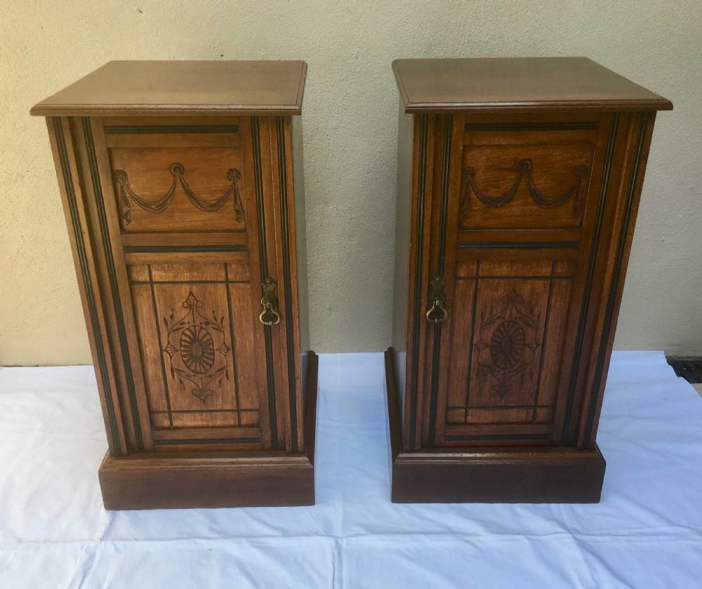 A matching pair of slim Edwardian mahogany bedside cabinets featuring carved swag decoration, single cupboards, brass handles, and solid plinth bases.