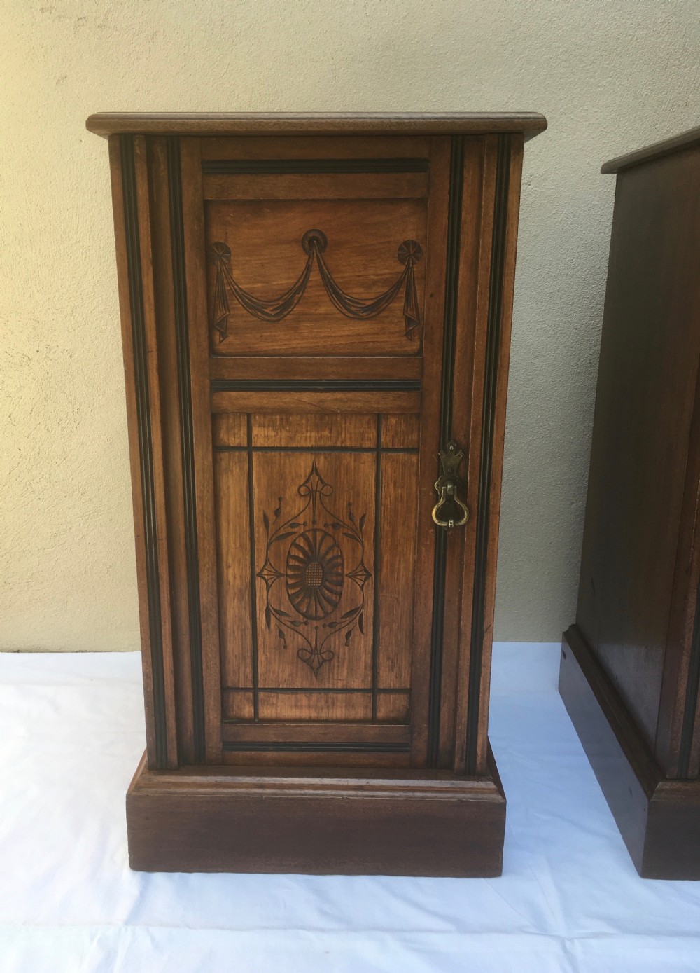 A matching pair of slim Edwardian mahogany bedside cabinets featuring carved swag decoration, single cupboards, brass handles, and solid plinth bases. View V