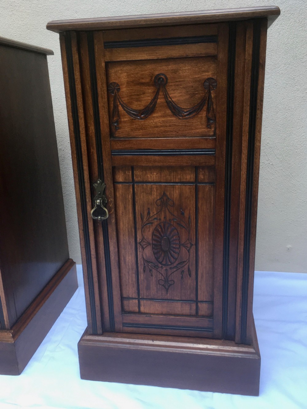 A matching pair of slim Edwardian mahogany bedside cabinets featuring carved swag decoration, single cupboards, brass handles, and solid plinth bases. View VI