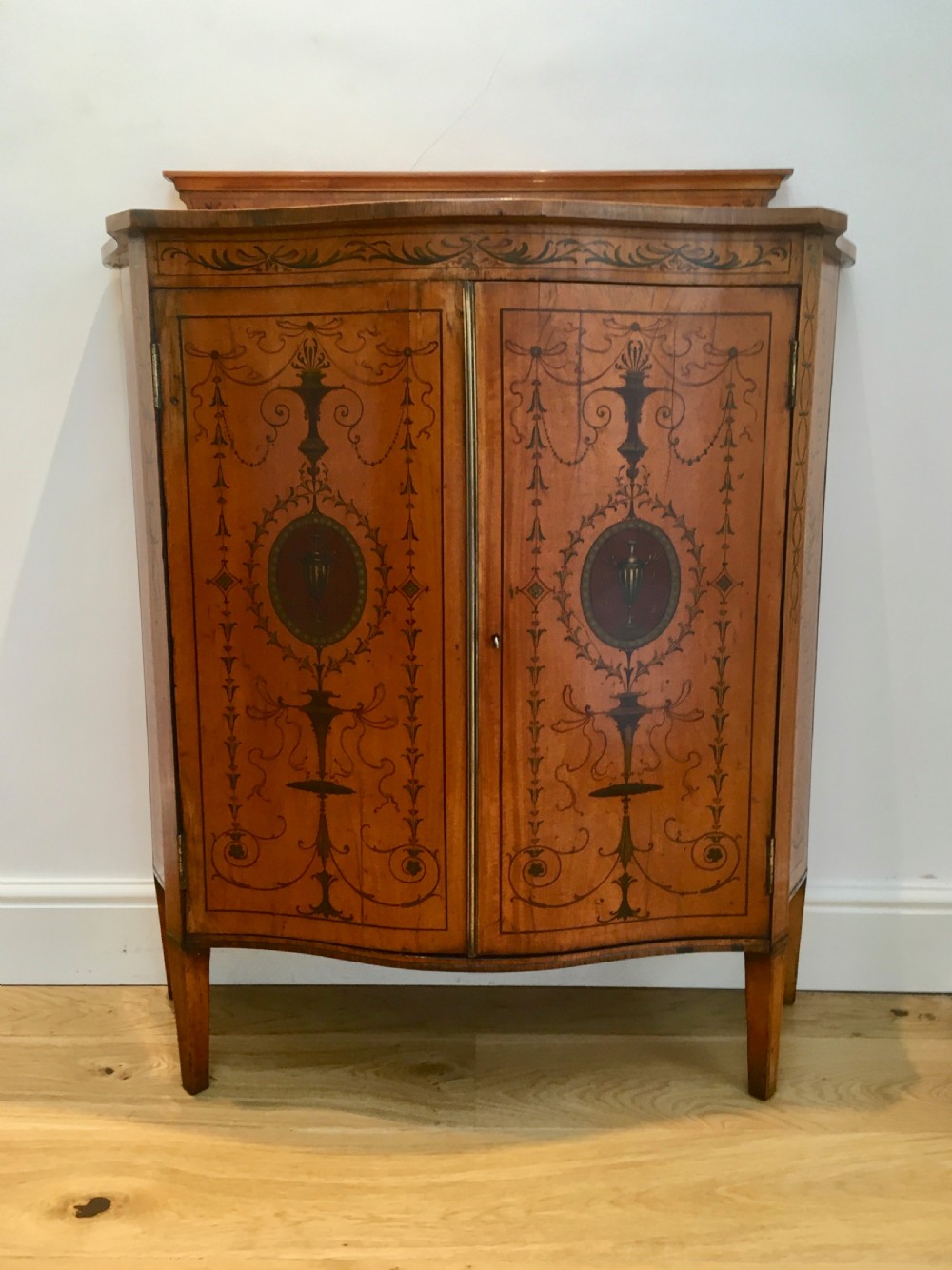 A satinwood side cabinet featuring a serpentine front, twin cupboard doors decorated with hand-painted Neoclassical urns and floral swags, supported by tapered legs.