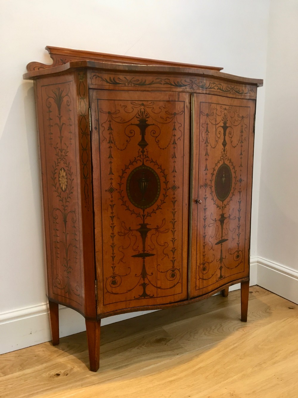 A satinwood side cabinet featuring a serpentine front, twin cupboard doors decorated with hand-painted Neoclassical urns and floral swags, supported by tapered legs. View II
