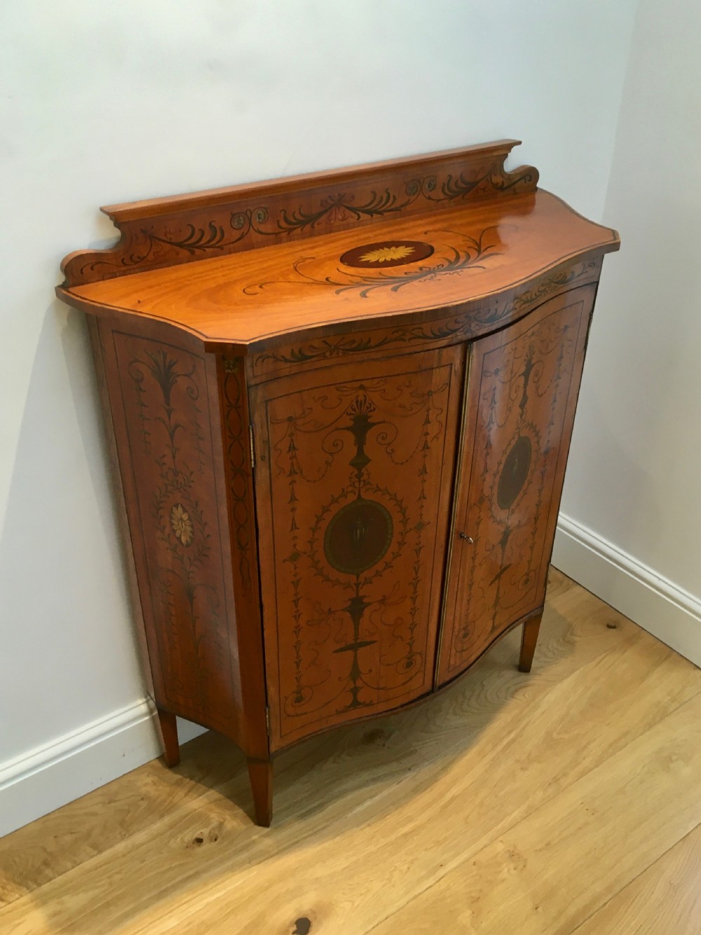 A satinwood side cabinet featuring a serpentine front, twin cupboard doors decorated with hand-painted Neoclassical urns and floral swags, supported by tapered legs. View III