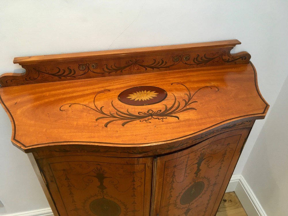 A satinwood side cabinet featuring a serpentine front, twin cupboard doors decorated with hand-painted Neoclassical urns and floral swags, supported by tapered legs. View IV