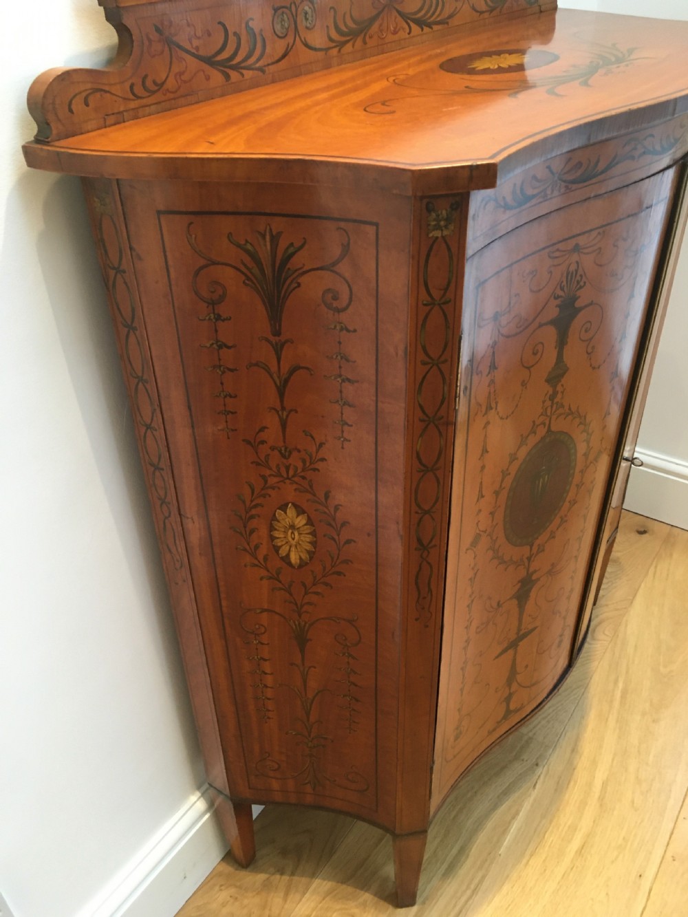 A satinwood side cabinet featuring a serpentine front, twin cupboard doors decorated with hand-painted Neoclassical urns and floral swags, supported by tapered legs. View V