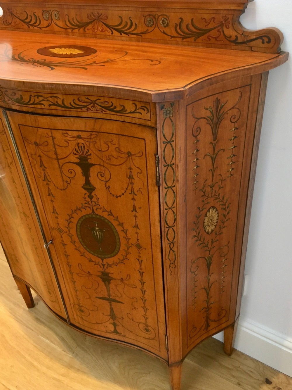 A satinwood side cabinet featuring a serpentine front, twin cupboard doors decorated with hand-painted Neoclassical urns and floral swags, supported by tapered legs. View VI