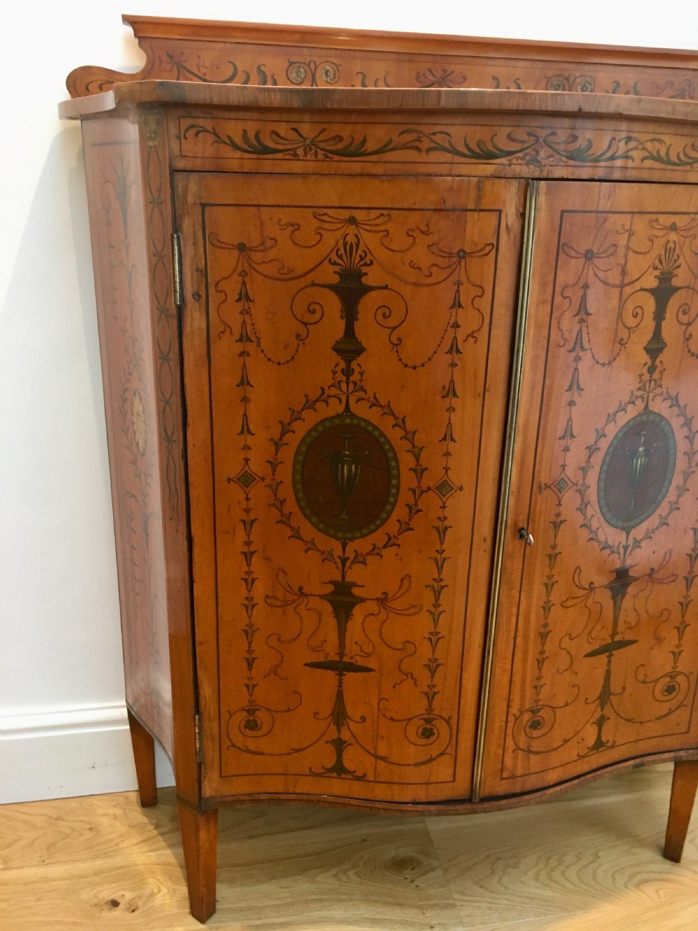 A satinwood side cabinet featuring a serpentine front, twin cupboard doors decorated with hand-painted Neoclassical urns and floral swags, supported by tapered legs. View VII