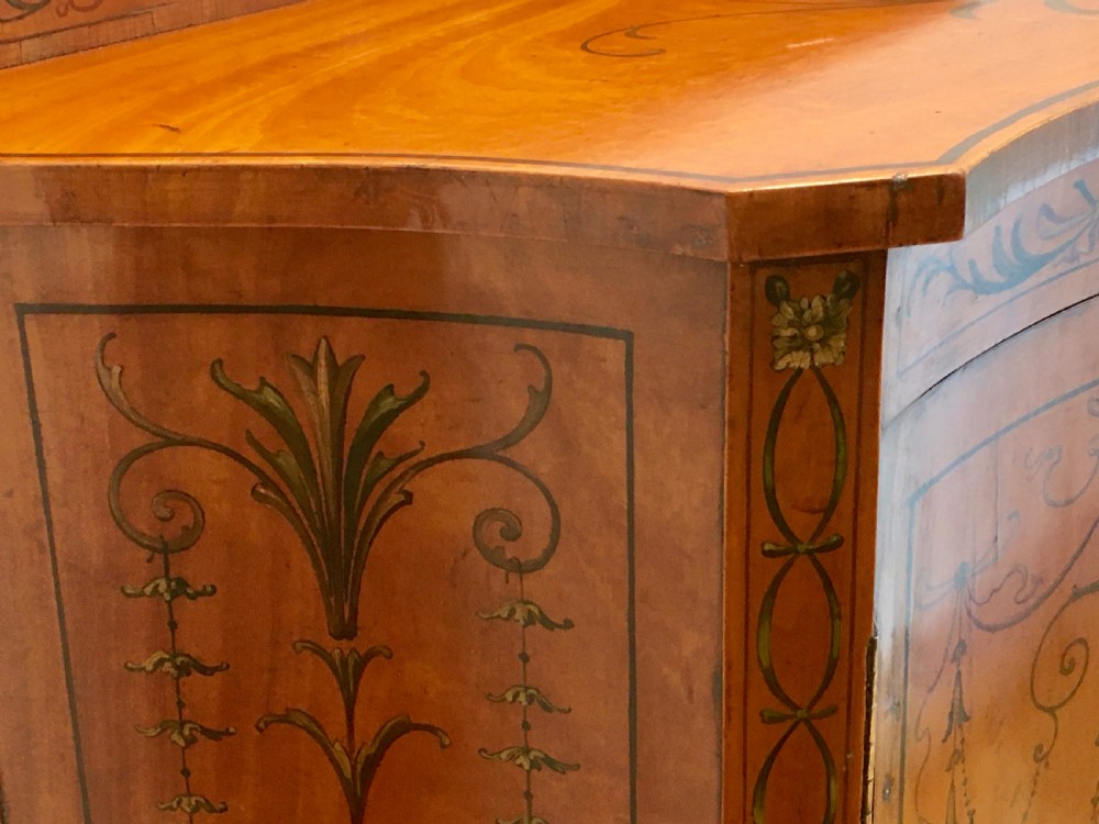 A satinwood side cabinet featuring a serpentine front, twin cupboard doors decorated with hand-painted Neoclassical urns and floral swags, supported by tapered legs. View XIV